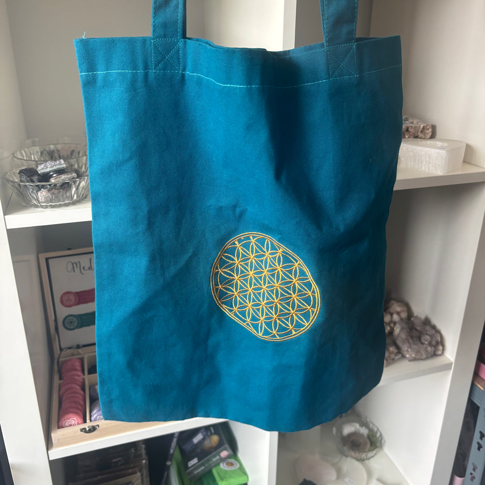 A teal blue canvas tote bag with a yellow geometric flower-of-life design printed on the front, hanging on a white shelf.
