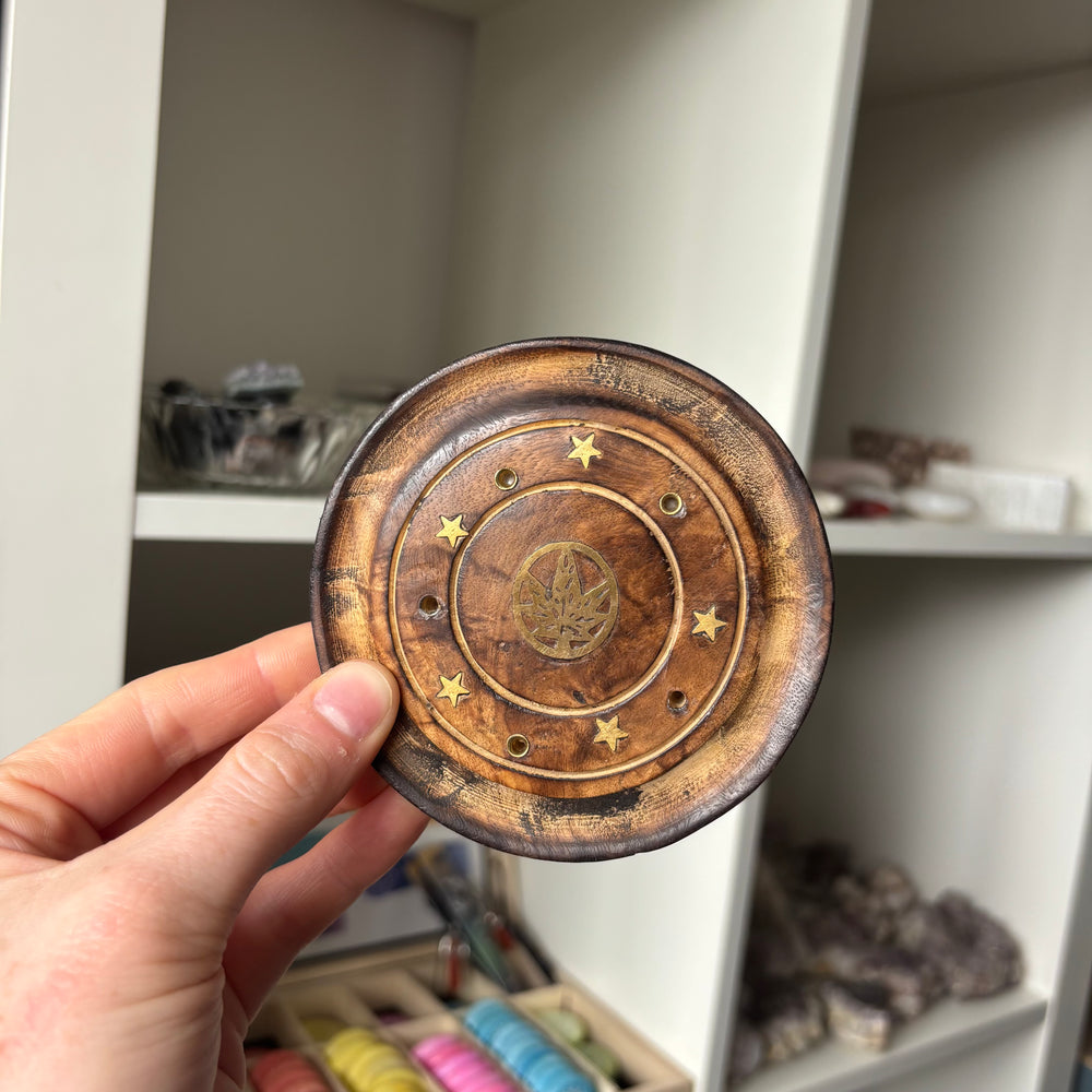 A close-up of a round wooden tray or coaster being held by a hand. The wood has a dark, distressed finish with concentric circular grooves, small star-shaped brass inlays, and a central leaf emblem.