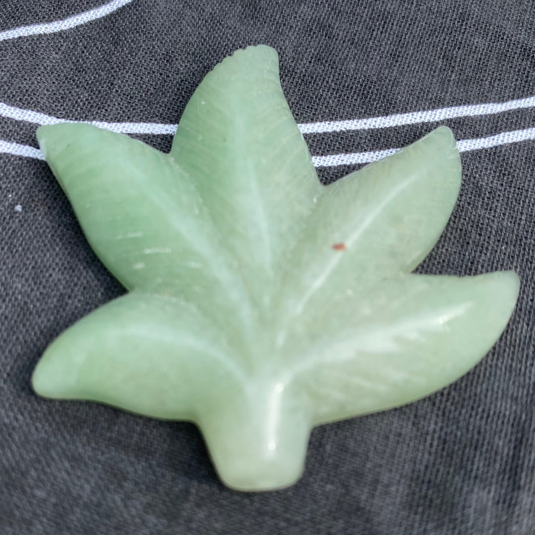 A light green jadeite leaf-shaped pendant with detailed carvings, resting on a dark gray textured fabric with white geometric patterns.