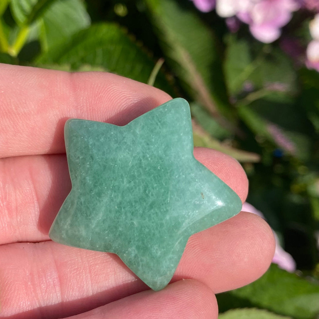A green star-shaped polished stone held in a person's hand, with blurred green leaves and pink flowers in the background.