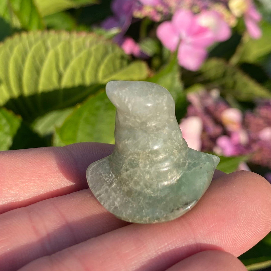 A small polished light green stone figurine shaped like a hat, held in a person's hand with a blurred garden background of green leaves and pink flowers.