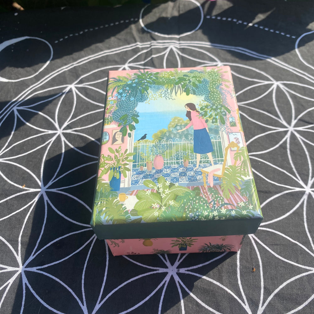 A rectangular decorative box with a colorful illustrated lid showing a woman in a garden, placed on a black fabric surface with a white geometric pattern.