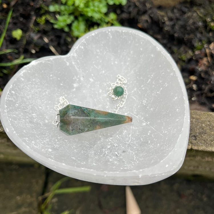 A heart-shaped green moss agate crystal pendulum with a silver chain, placed on a heart-shaped stone on a bright sunny day.