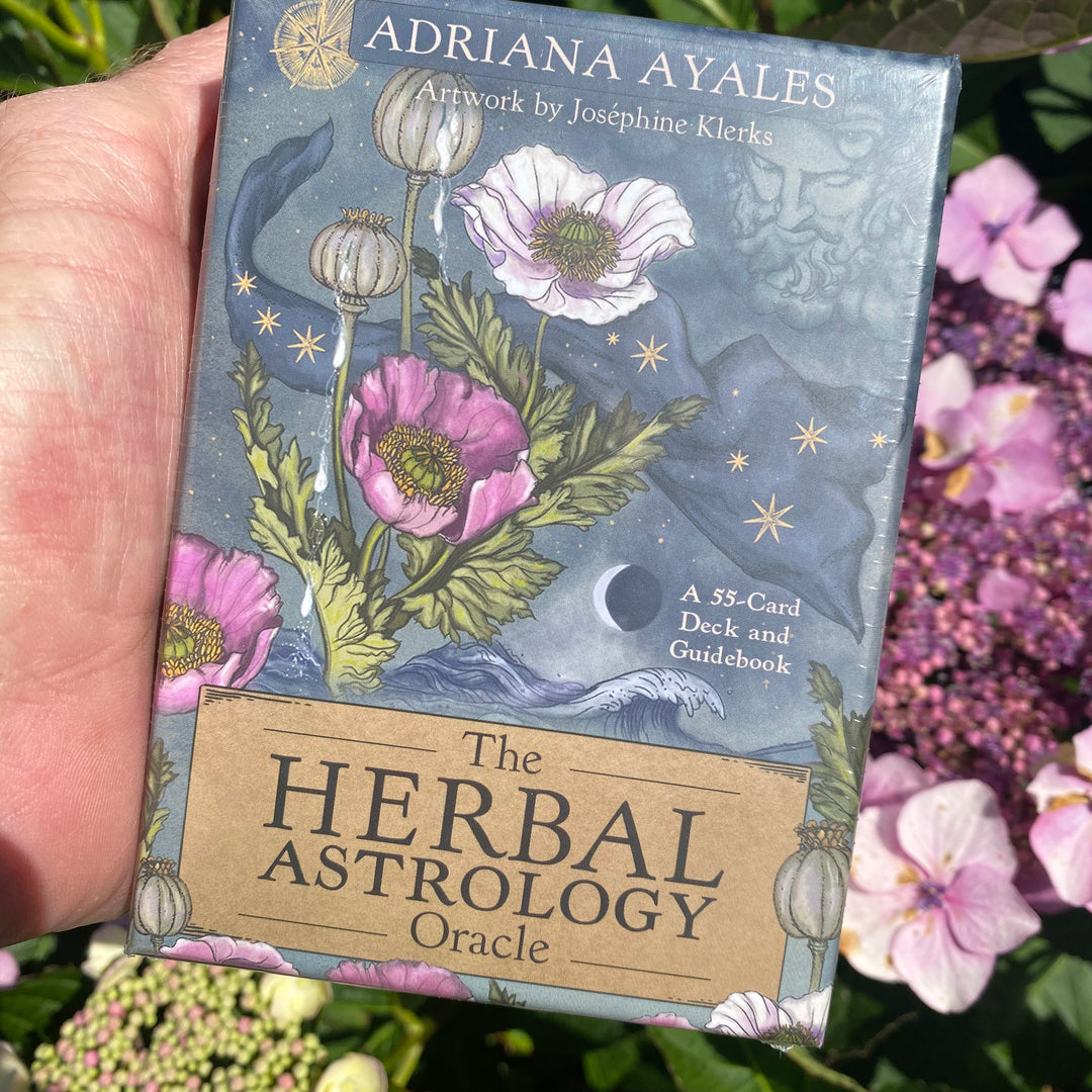 A hand holds 'The Herbal Astrology Oracle' deck and guidebook box by Adriana Ayales. The box features a dark blue night-sky and moon illustration with pink poppy flowers and green leaves, set against a background of blurred pink flowers.
