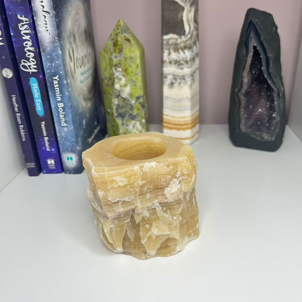 A beige honey calcite tea light holder placed on a shelf with various crystal decorations and books in the background.