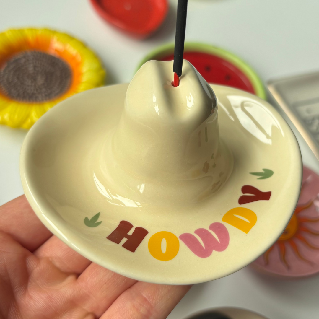 Ceramic incense holder with 'Howdy' text held by a hand, with colorful ceramic discs in the background.