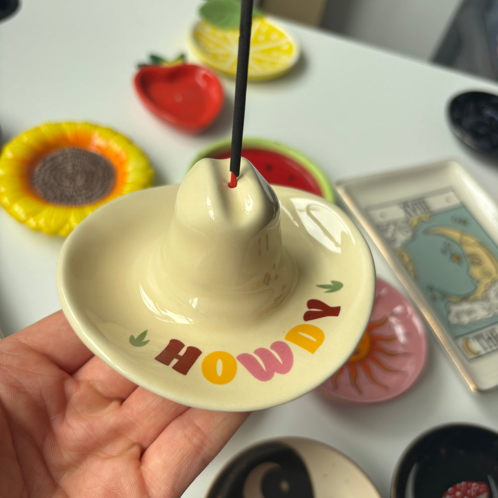 A hand holds a ceramic incense holder shaped like a sombrero with the word 'HOWDY' painted on the front. A black incense stick is inserted into the holder.