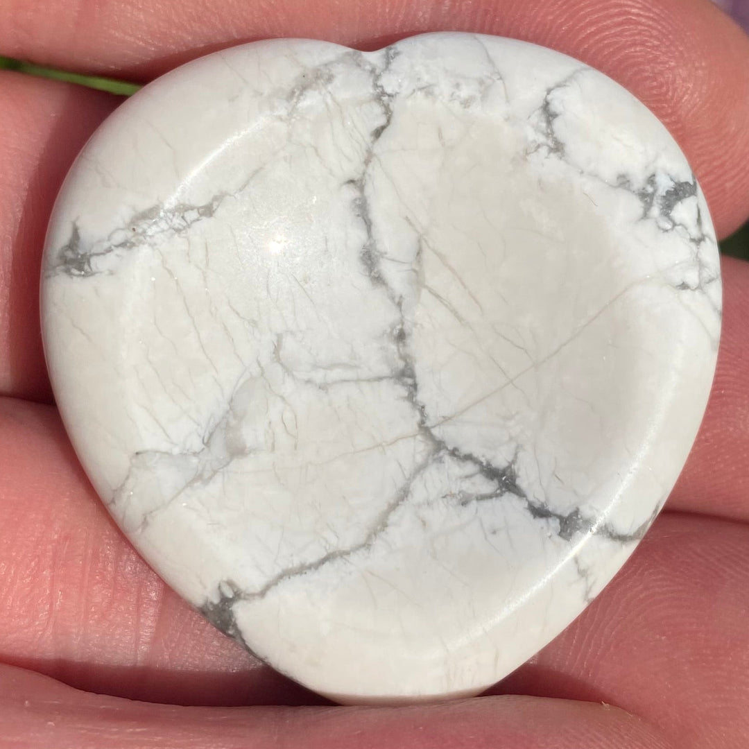 A heart-shaped Howlite crystal with gray veining, held in a person's hand with blurred pink flowers and green foliage in the background.