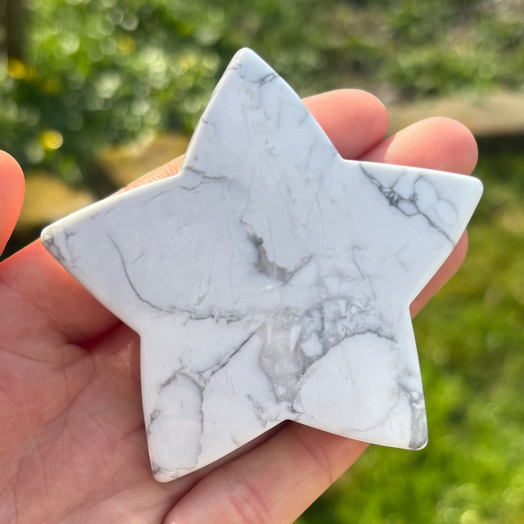A hand holding a large white howlite star crystal carving against a blurred background of a garden.