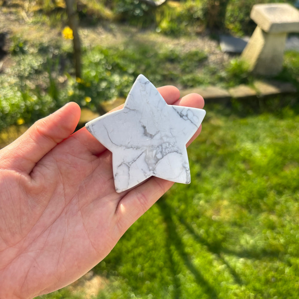 A hand holding a large white howlite star crystal carving against a blurred background of a garden.