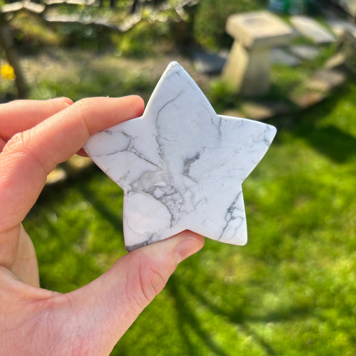 A hand holding a large white howlite star crystal carving against a blurred background of a garden.
