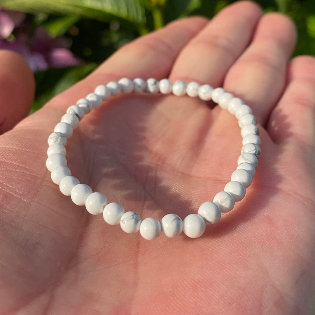 A white beaded Howlite Mini Round Beaded Bracelet resting on an open hand, with green leaves and purple flowers blurred in the background.