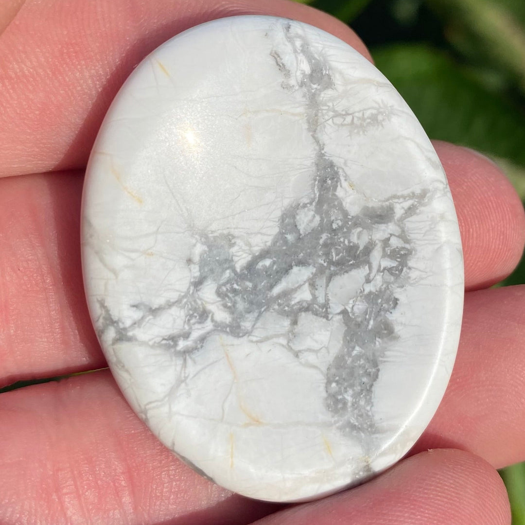 A polished white Howlite Oval Worry Stone with gray and subtle gold veining, held in a person's hand against a background of green leaves and pink flowers.