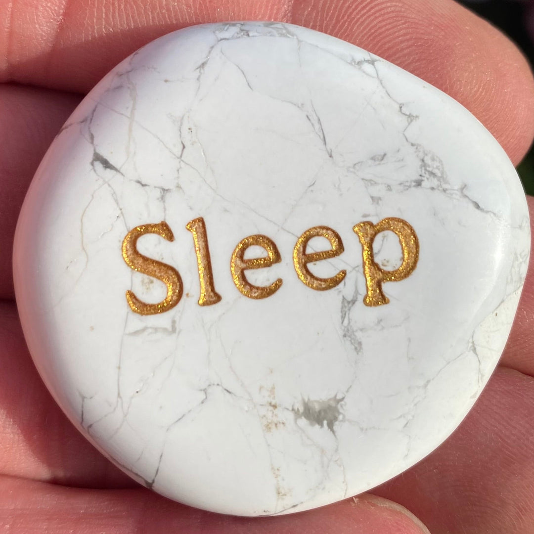 A white Howlite Smoothstone with the word 'Sleep' in gold foil, held in a person's hand.