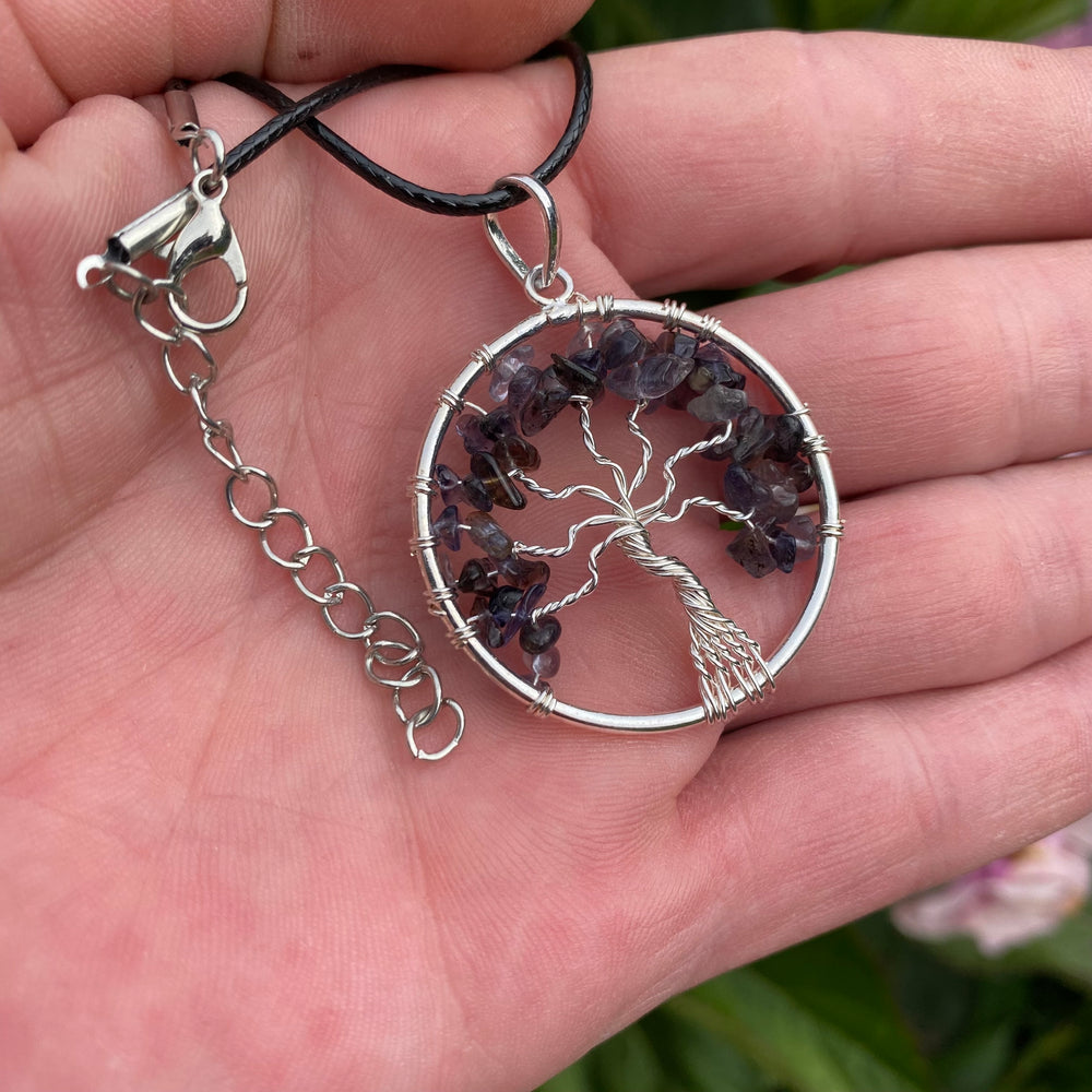 A silver tree of life pendant necklace with purple crystal accents, held in a person's hand. The pendant is suspended from a black cord with a silver lobster clasp and extension chain.