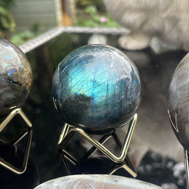 A polished labradorite crystal sphere with a blue iridescent flash, displayed on a gold metal stand.