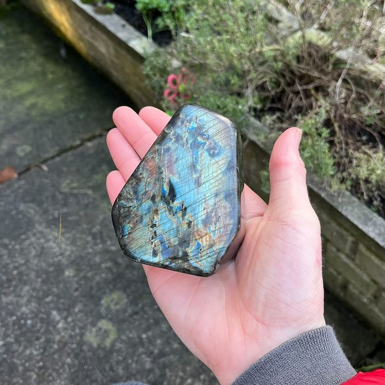 A hand holding a polished labradorite stone with a shimmering blue, green, and brown iridescent finish, set against an outdoor background of foliage and stone.