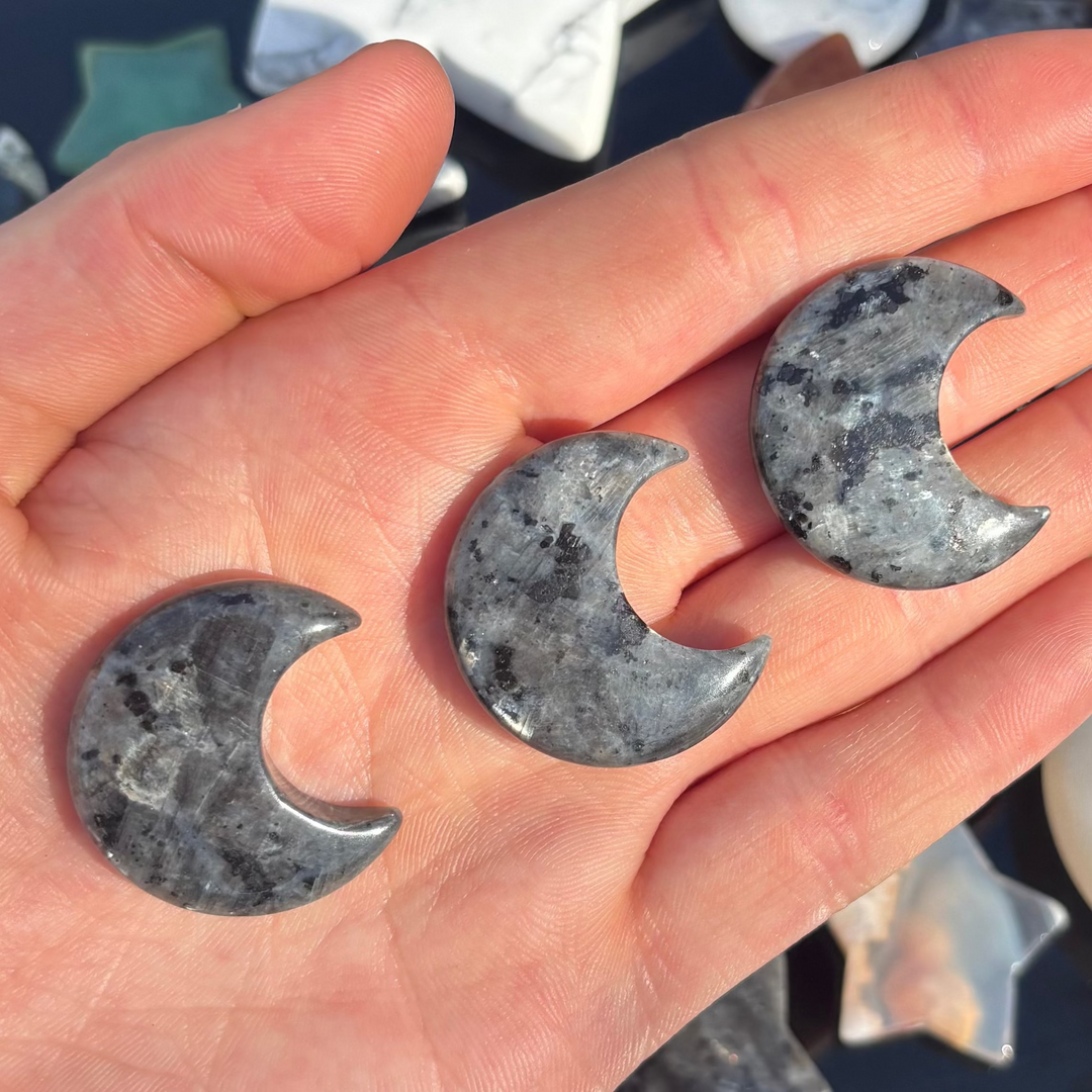 A hand holding three larvikite half moon shaped crystal carvings, with more carvings in various shapes arranged in the background.