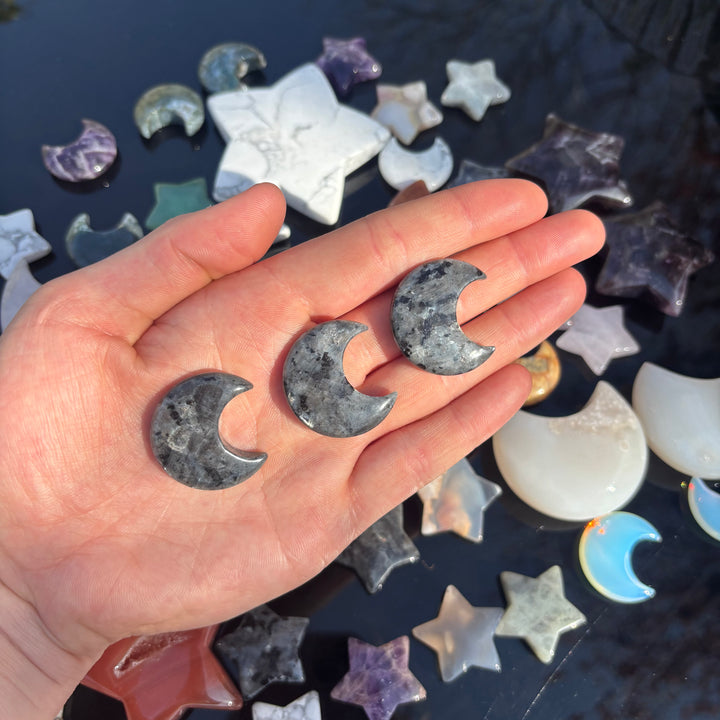 A hand holding three larvikite half moon shaped crystal carvings, with more carvings in various shapes arranged in the background.