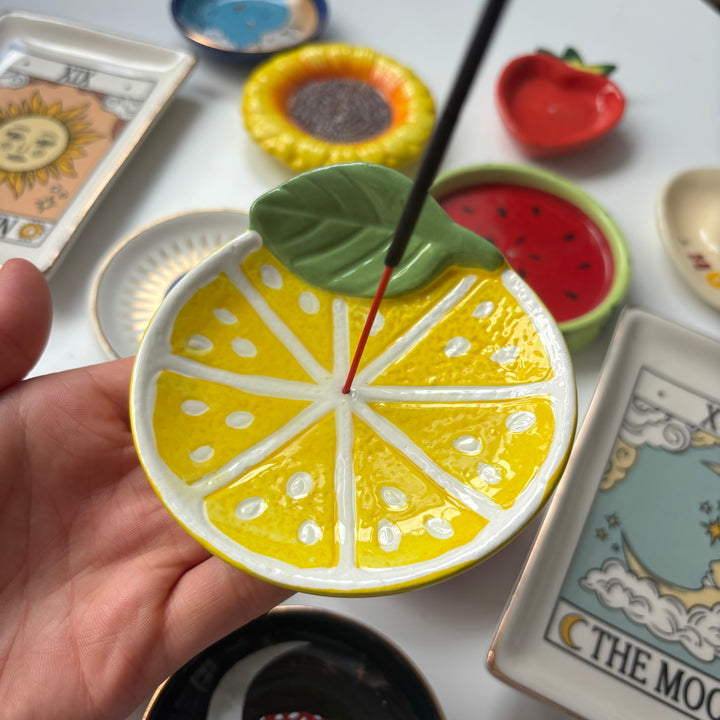 Hand holding a ceramic Lemon Incense Stick Holder with incense, surrounded by various small dishes and cards.