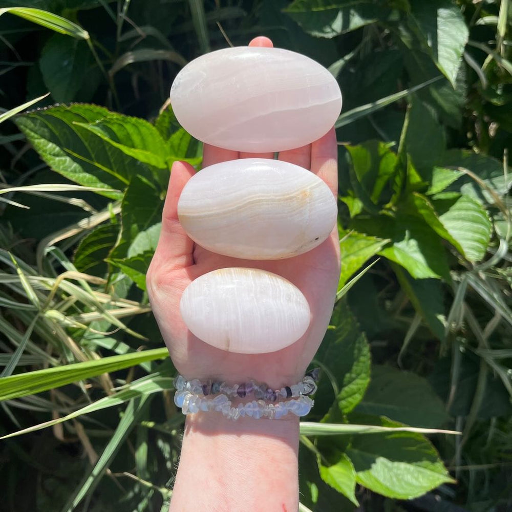 Three smooth, polished Mangano Calcite Palm Stones with subtle natural banding are held in a person's hand against a background of green outdoor foliage.
