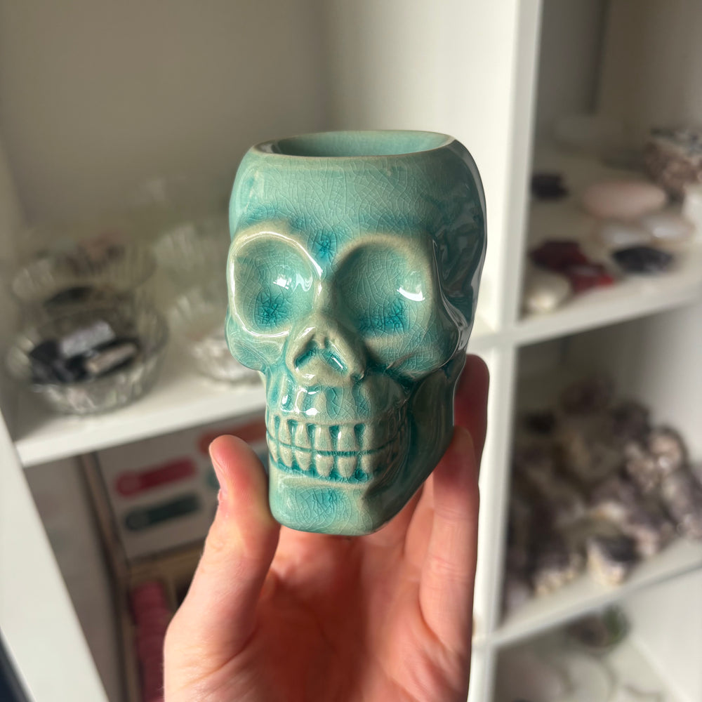 A Teal ceramic skull-shaped vase held in front of a shelf with various items.