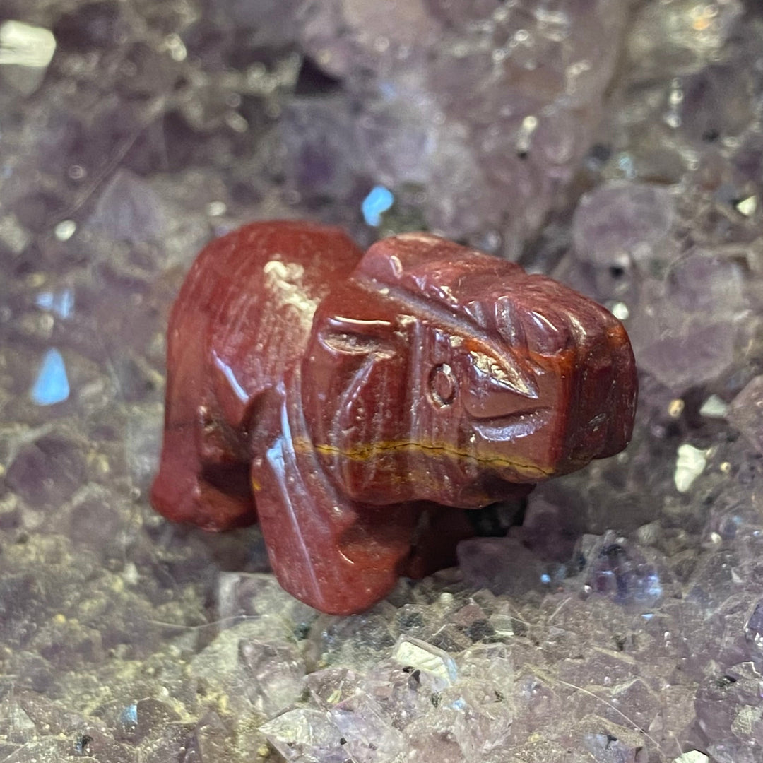 A small carved Mookaite Elephant Carving placed on a bed of purple and clear crystal clusters.