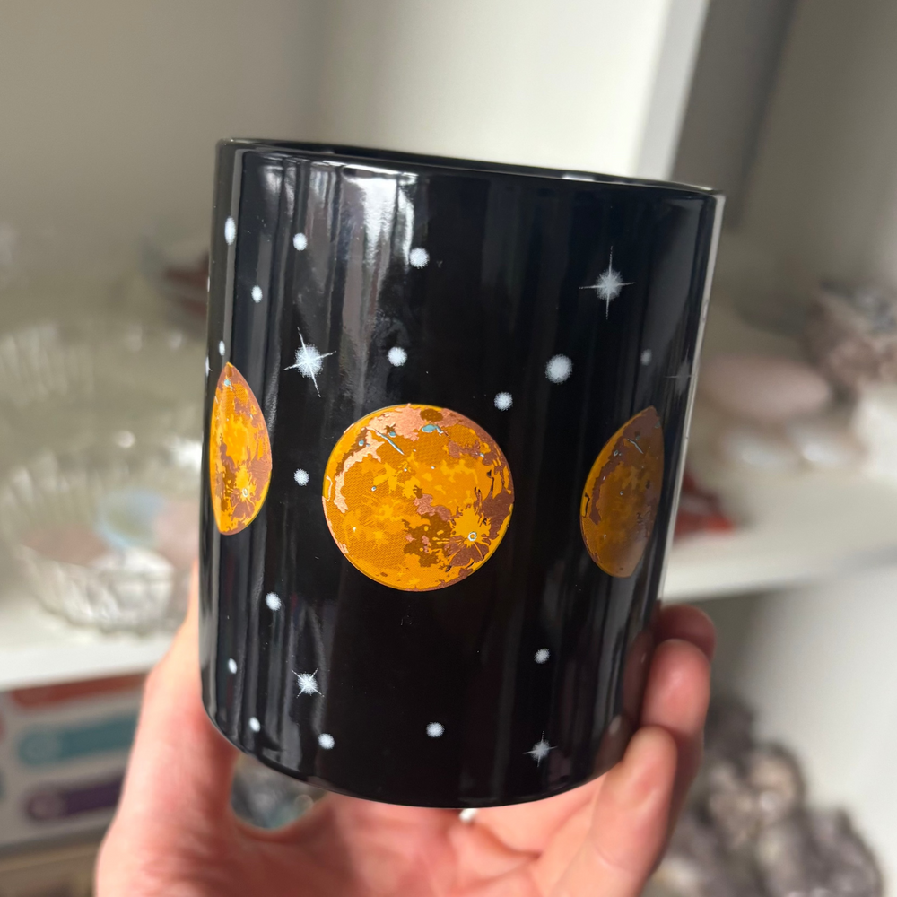 Black mug with orange moon phases held in a hand against a blurred indoor background