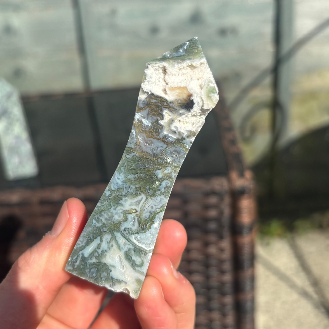 A hand holding a polished Moss Agate crystal tower with a blurred background.