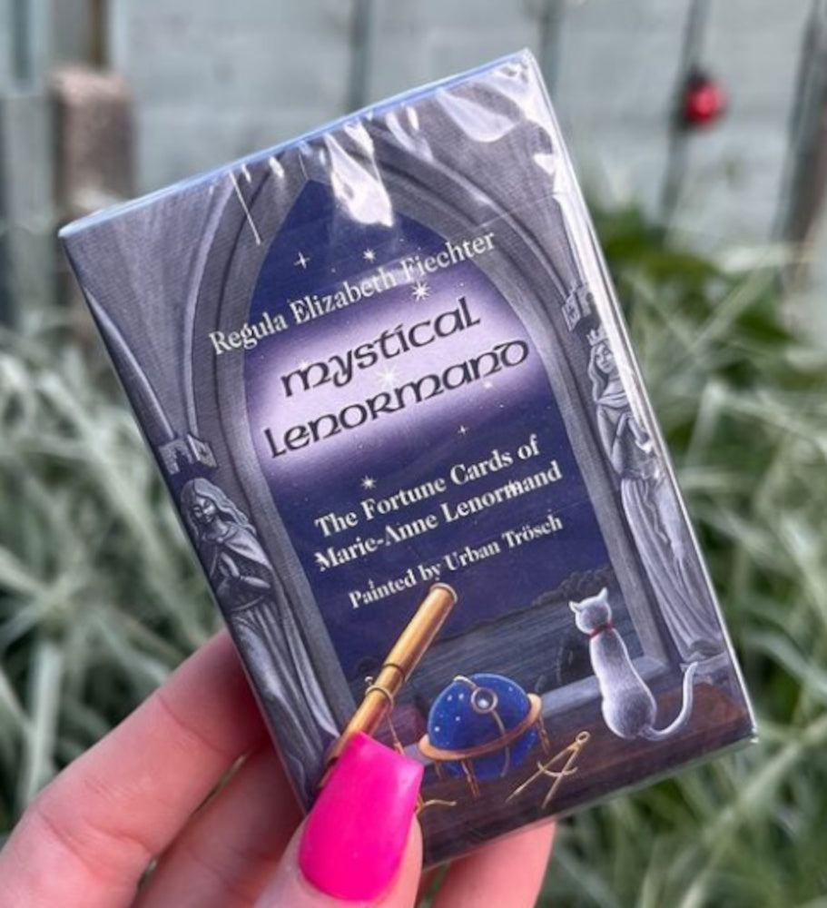 A hand holding a pack of Mystical Lenormand Mini Oracle Cards by Urban Trosch in a brightly lit garden