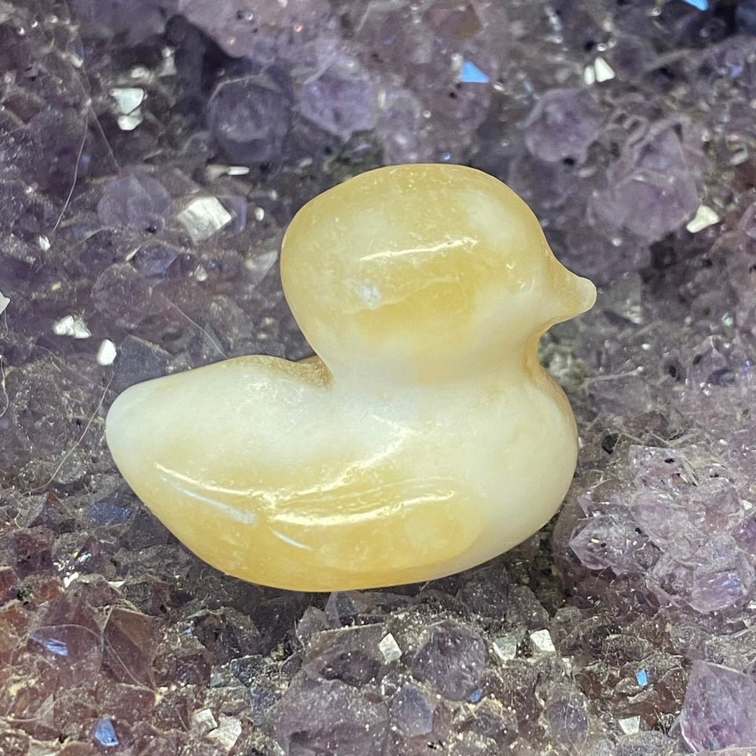 A small carved Orange Calcite Duck Carving resting on a bed of purple and brown crystal rocks.