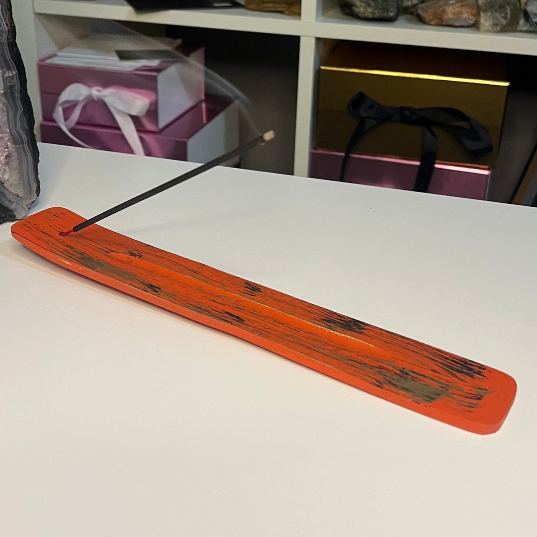 An orange painted incense stick holder made of wood, displayed on a white surface with various items in the background, including a crystal and some decorative boxes.