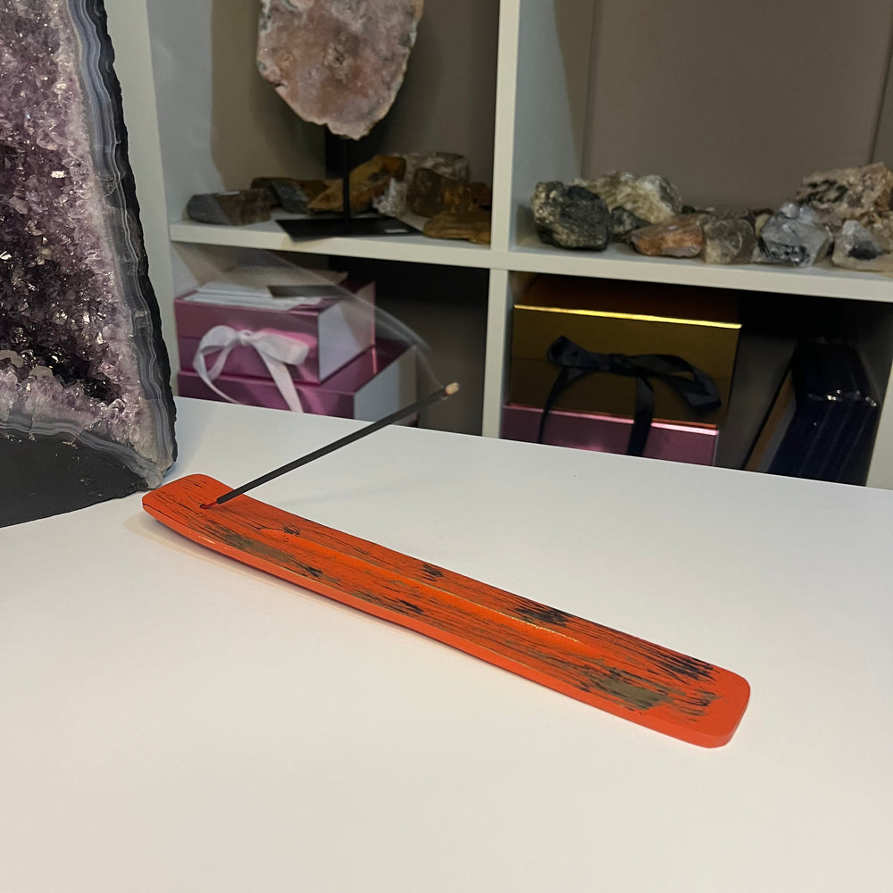 A rectangular orange incense holder with dark wood grain and a single incense stick resting diagonally across it, placed on a white tabletop with shelves of crystals and gift boxes in the background.