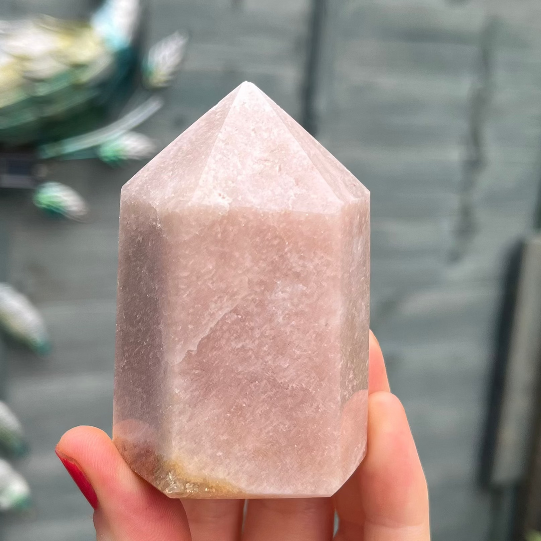 A hand holding a faceted rose quartz crystal tower against a blurred gray background with other crystals visible.
