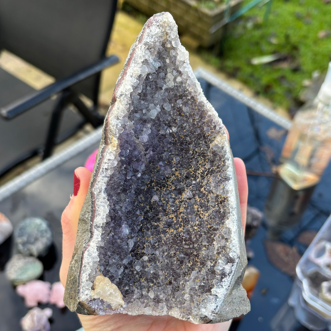 Hand holding a large crystal or mineral specimen outdoors with a chair and plants in the background.