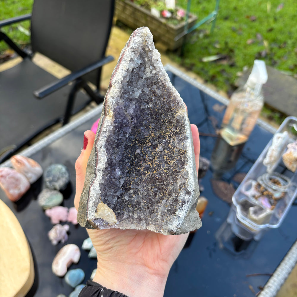 Hand holding a large crystal or mineral specimen outdoors with a chair and plants in the background on a bright sunny day.