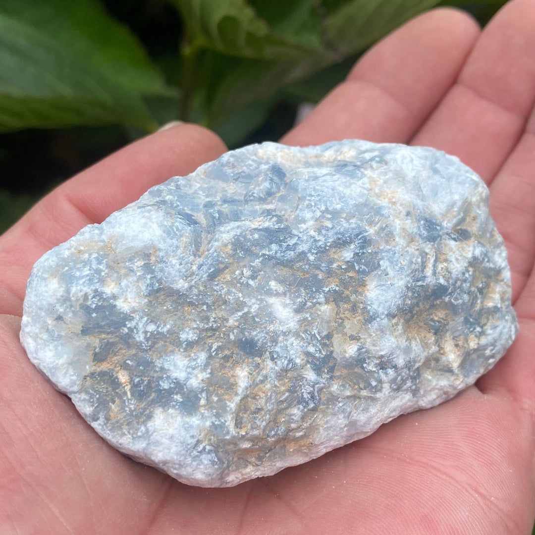 A chunk of Raw Blue Calcite Crystal held in a hand in a garden on a bright sunny day