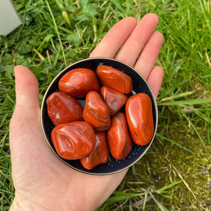 A hand holding a small black bowl filled with polished Red Jasper Tumble Stones, photographed outdoors against green grass.