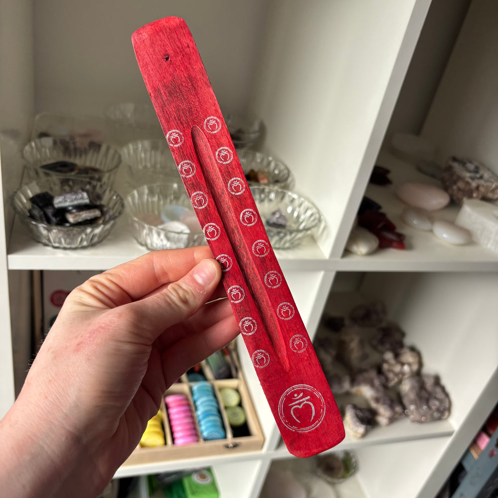 Red Root Chakra Incense Stick Holder with white patterns held by a hand in front of a shelf with various items.