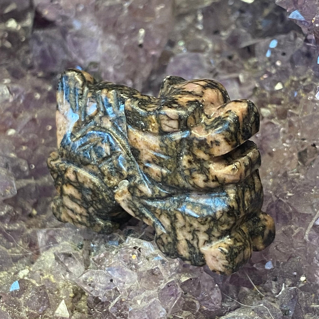 A Rhodonite Dragon Carving, with a tan and dark brown marbled pattern and subtle blue-green veining, displayed among purple crystal formations.