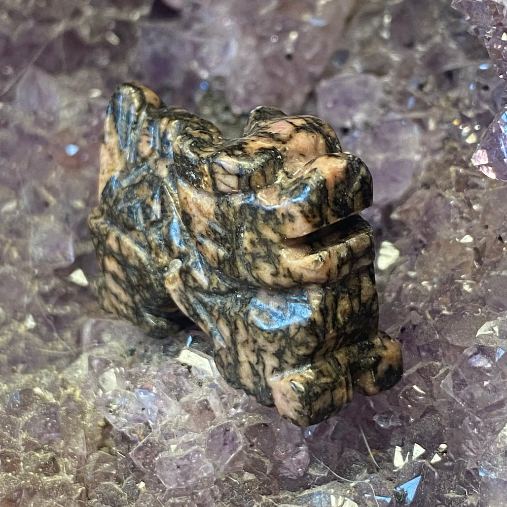 A small Rhodonite Dragon Carving with a mottled brown, black, and beige marbled pattern, resting on a purple crystal cluster.