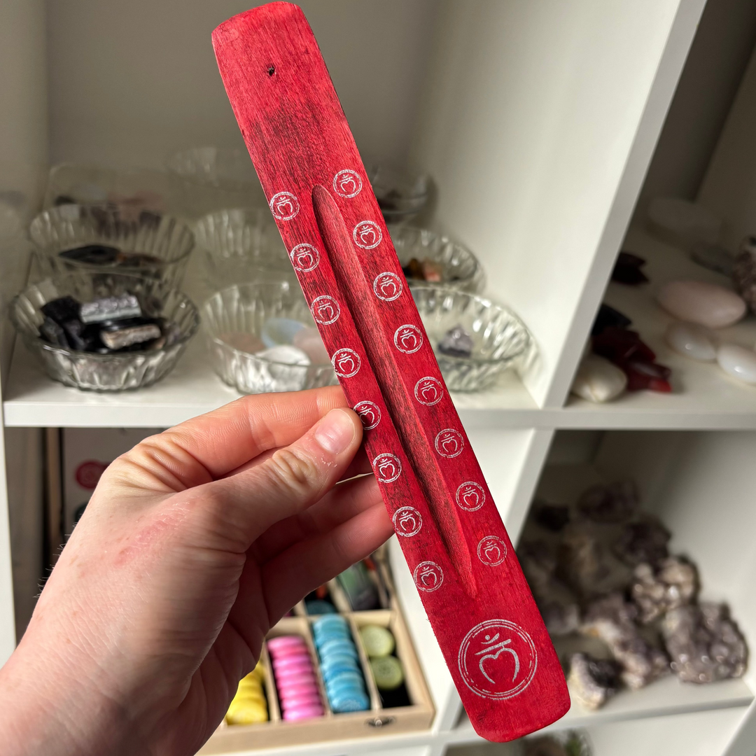 Red Root Chakra Incense Stick Holder with white patterns held by a hand in front of a shelf with various items.
