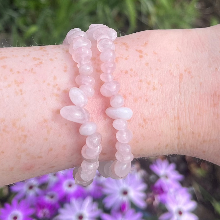 A person is wearing a bracelet made of rose quartz crystal chips, displayed on a wrist with a background of plants and flowers.