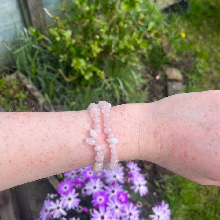 A person is wearing a bracelet made of rose quartz crystal chips, displayed on a wrist with a background of plants and flowers.