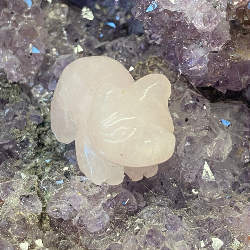 A small Rose Quartz Pig Carving resting on a bed of purple and lavender crystal clusters.