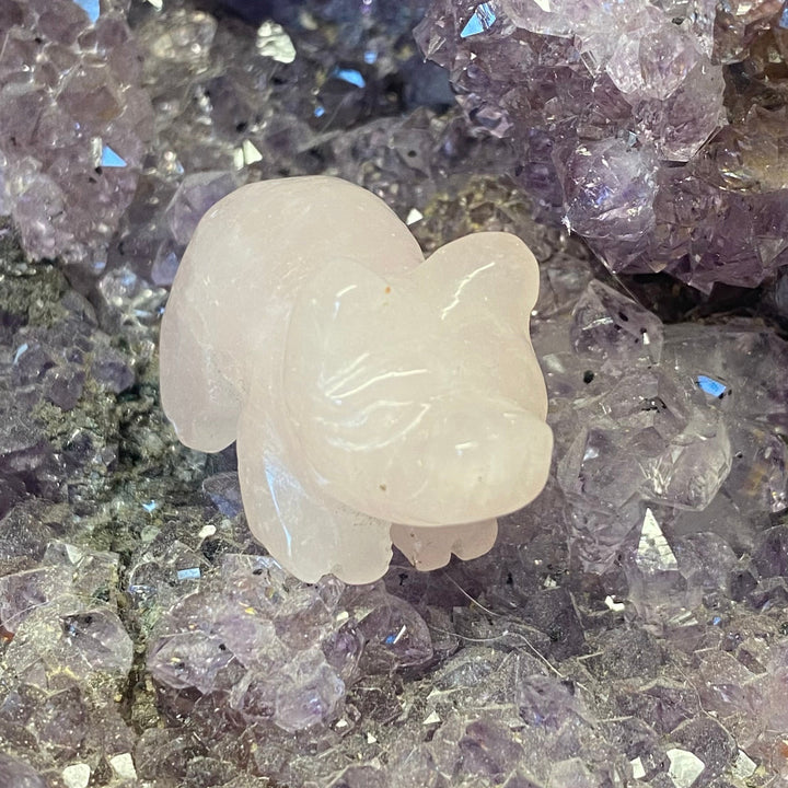 A small Rose Quartz Pig Carving resting on a bed of purple and lavender crystal clusters.