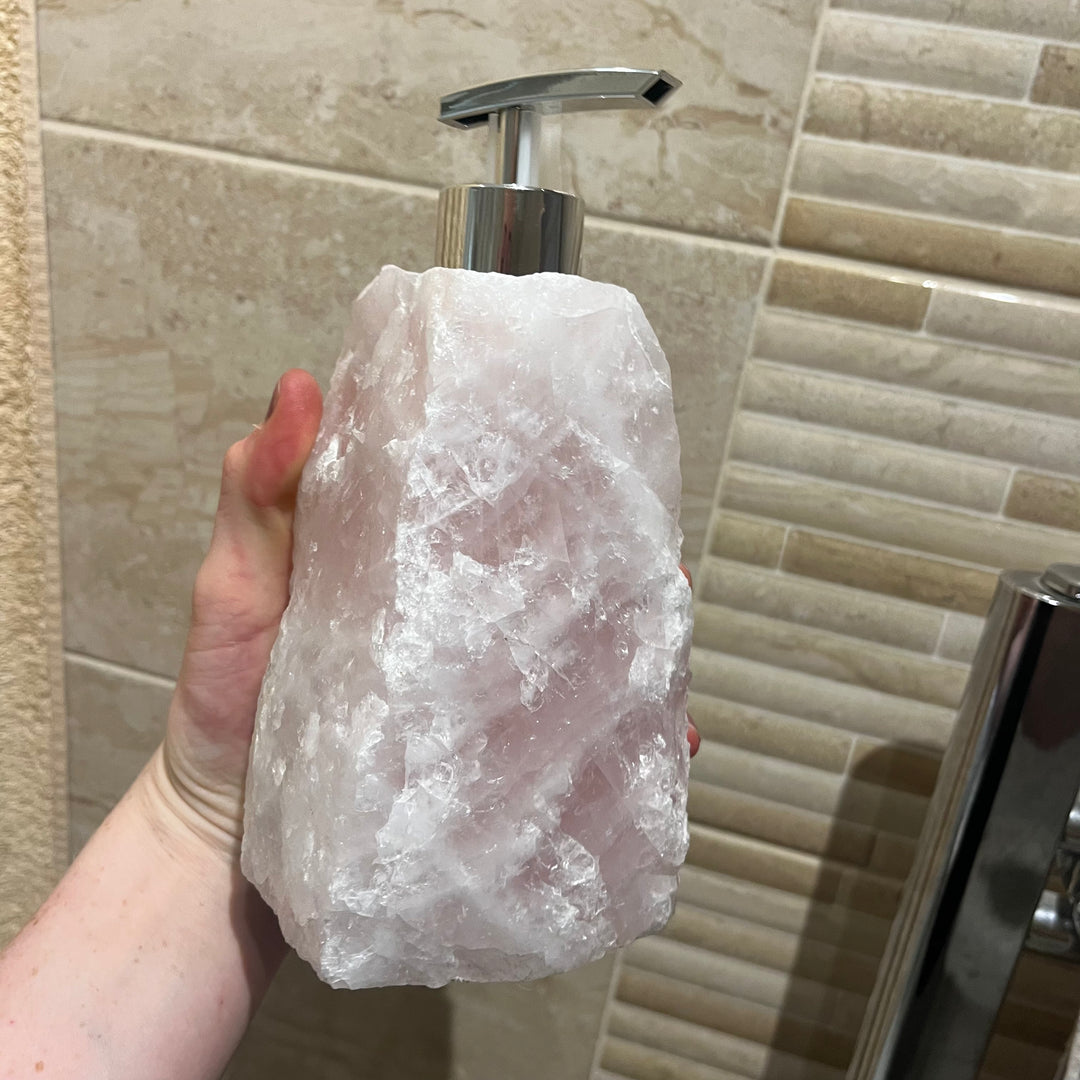 A substantial raw Rose Quartz crystal soap dispenser held up to view, showcasing its heavy block shape, soft icy-pink colour, and rugged unpolished stone texture topped with a metallic silver pump head.