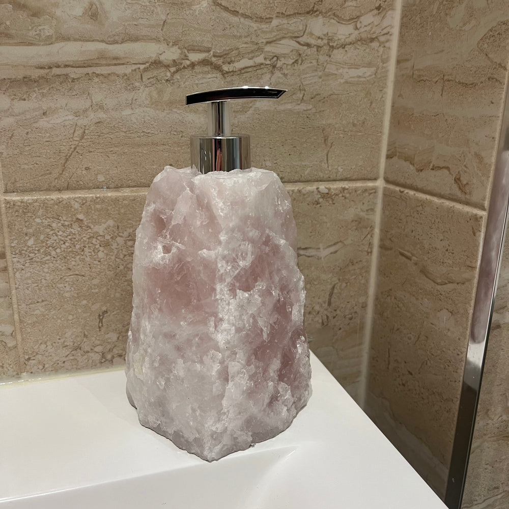 A raw pink Rose Quartz gemstone liquid soap holder resting elegantly on the white corner edge of a tiled bathroom sink basin.