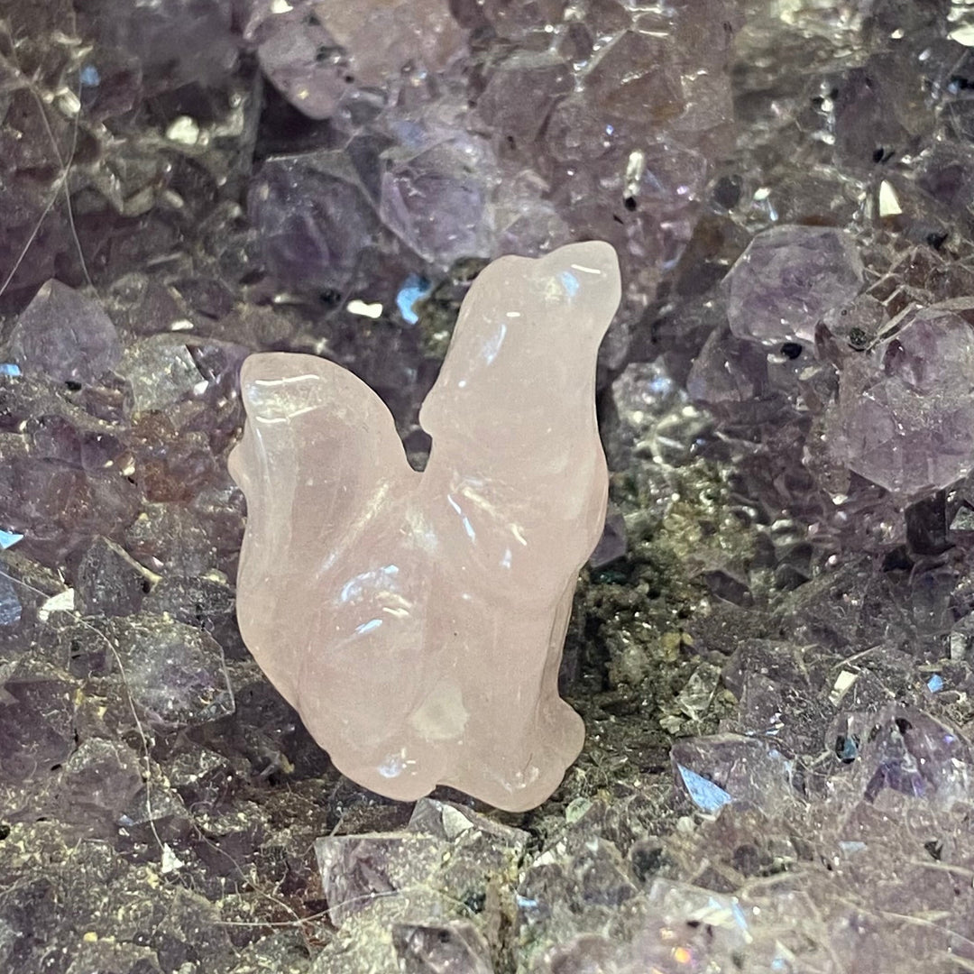 A small carved pale pink Rose Quartz Wolf Carving placed on a bed of purple and gray crystal clusters.