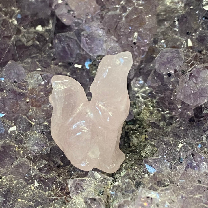 A small carved pale pink Rose Quartz Wolf Carving placed on a bed of purple and gray crystal clusters.